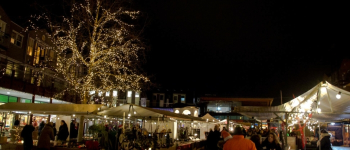 Een totaaloverzicht van de Kerstmarkt Westervoort. Veel marktkramen en kerstverlichting. Ook de boom is geheel verlicht met lampjes. Het is donker buiten, waardoor de kerstverlichting voor sfeer zorgt. Er lopen mensen langs de kramen. Kerst, Kerstmarkt, Westervoort, kerstverlichting, verlichting, lampjes, kramen, marktkramen. 